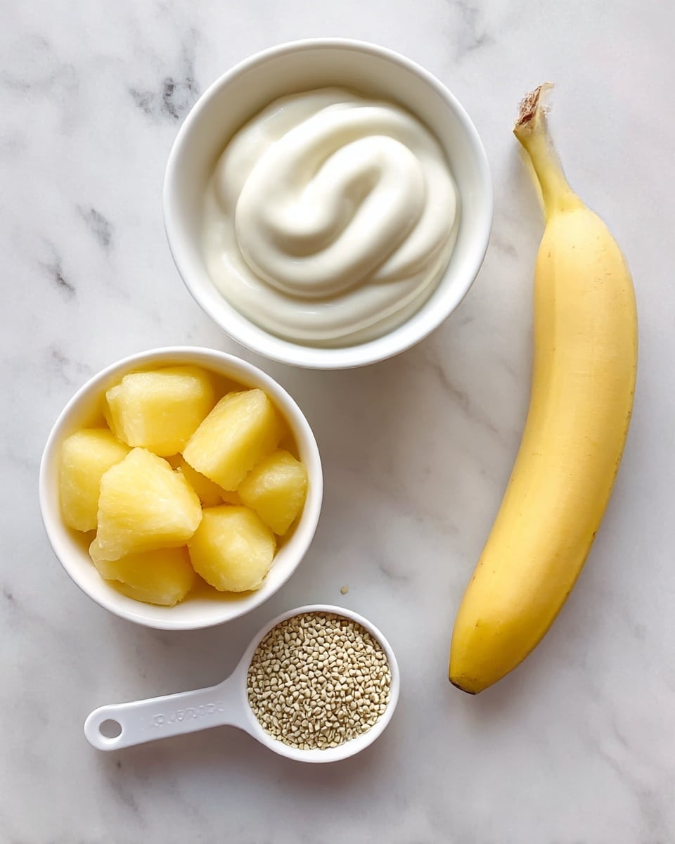 A white bowl filled with bright yellow frozen pineapple chunks sits on a white marbled surface next to a small whole yellow banana lying flat. Nearby, a white cup holds thick, creamy vanilla Greek yogurt with smooth swirls on top. A white measuring spoon filled with small, beige hemp hearts rests beside the yogurt. Each item is spaced clearly with natural light highlighting soft textures and fresh colors photo taken with an iphone --ar 4:5 --v 7