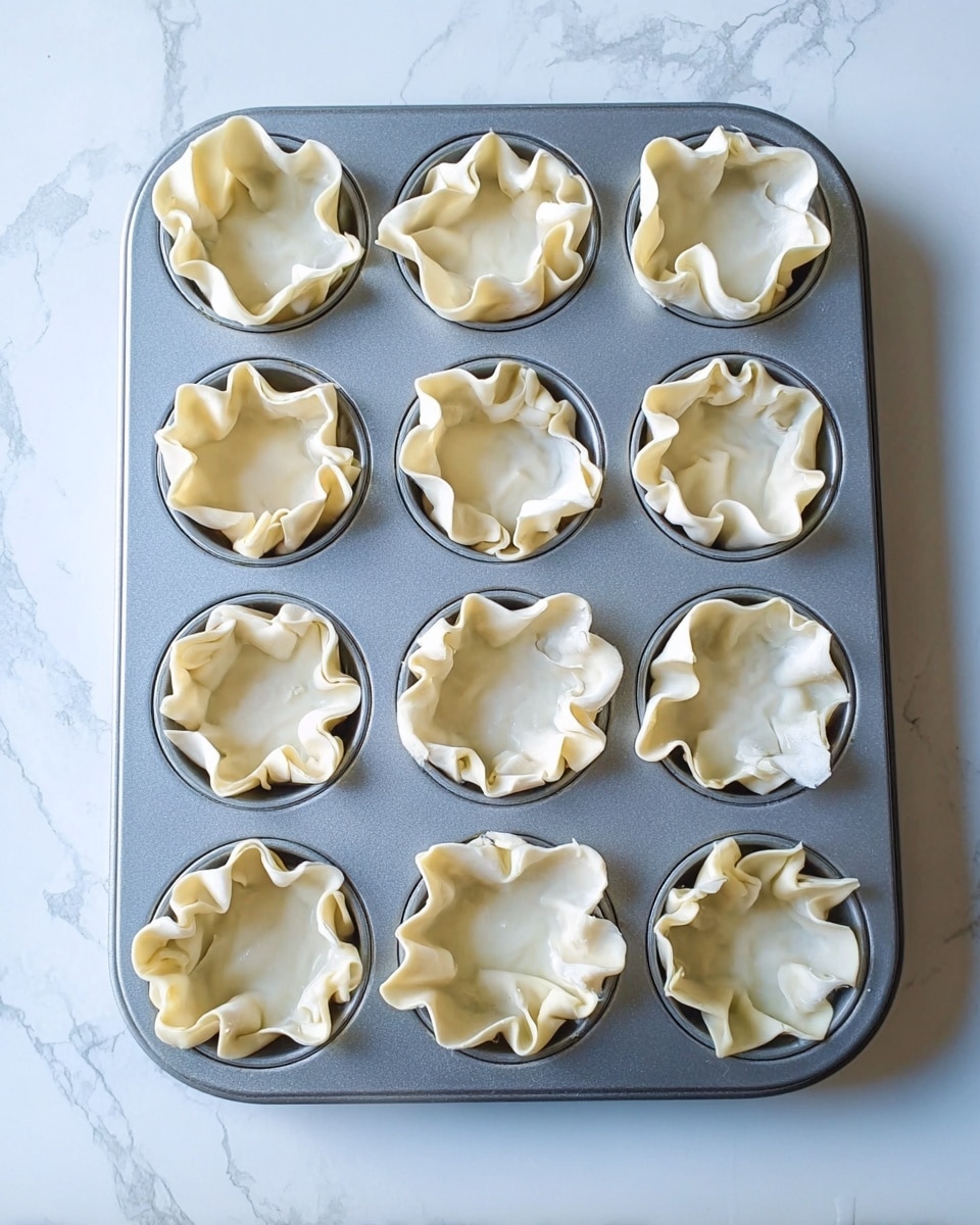 A grey metal muffin tray with 12 cups is placed on a white marbled surface. Each cup holds a single layer of pale, thin dough with soft folds and waves, forming a loosely ruffled cup shape. The dough edges rise above the rim of each muffin cup, creating a flower-like texture. The light highlights the smooth, slightly shiny surface of the dough, emphasizing the soft curves and thinness of each layer. photo taken with an iphone --ar 4:5 --v 7