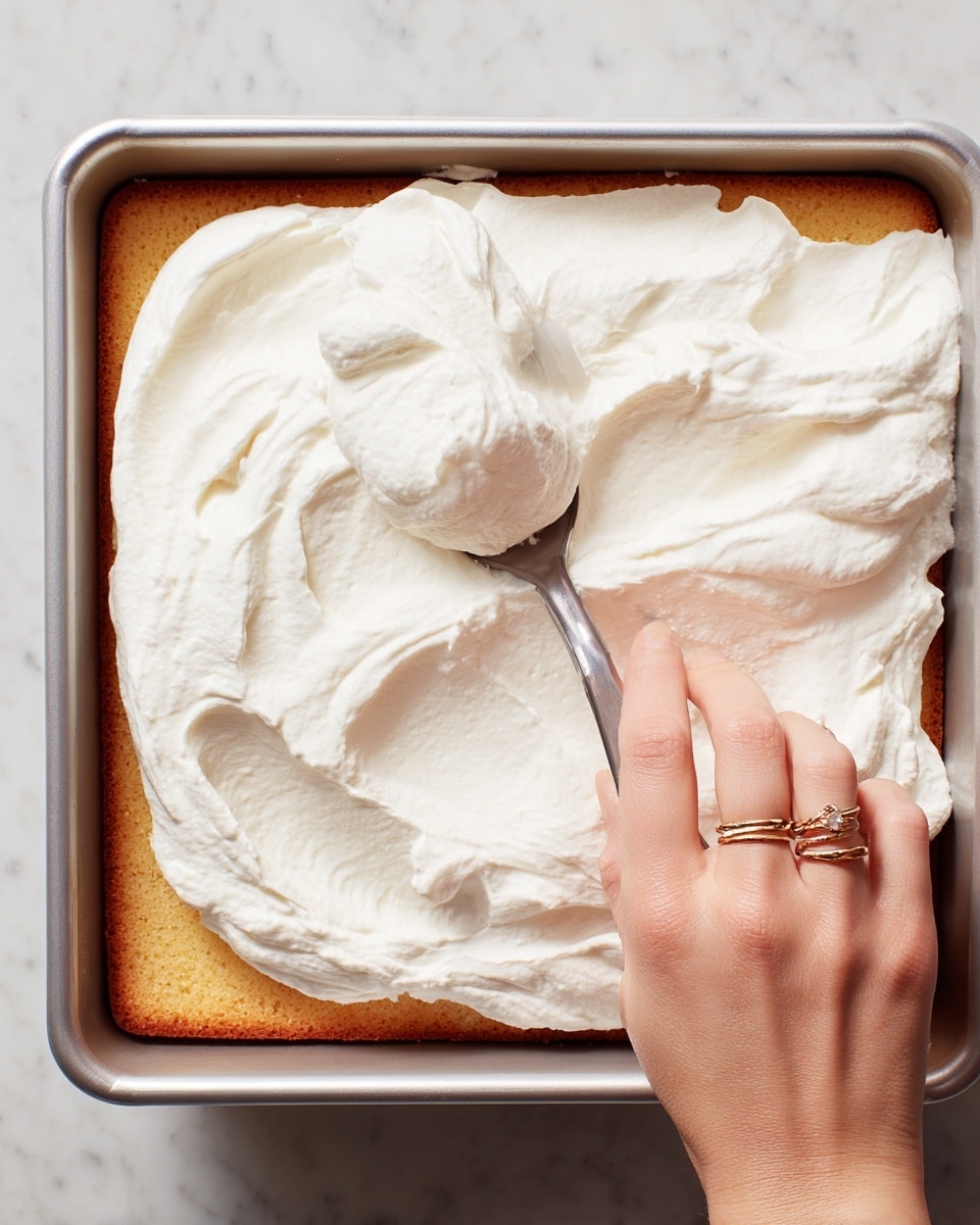 A close-up view of a square pan with a golden-brown cake layer at the bottom, covered halfway with thick, white whipped cream being spread gently by a woman's hand holding a silver spoon. The whipped cream has a smooth and fluffy texture with soft peaks, and the woman’s hand has two gold rings on the fingers. The pan is placed on a white marbled surface. Photo taken with an iphone --ar 4:5 --v 7