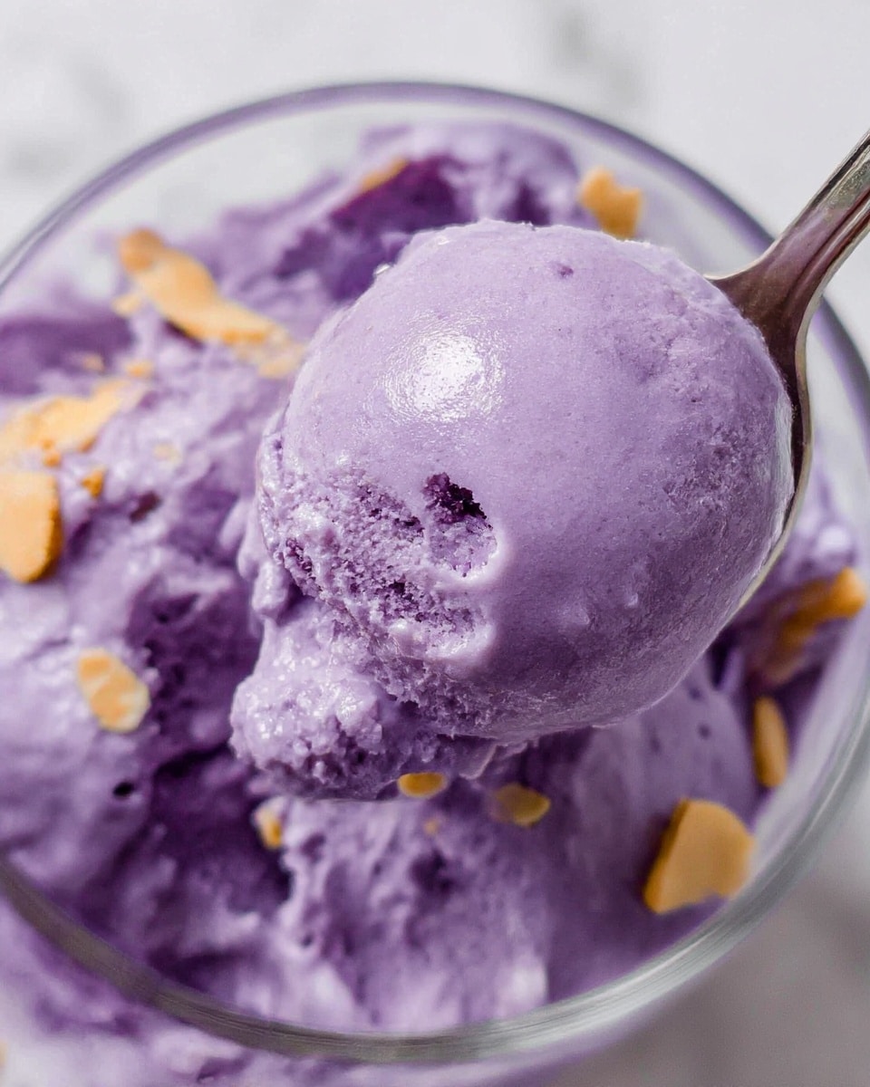 A close-up shot of a glass bowl filled with creamy, smooth purple ice cream. The ice cream has a rich, thick texture with some small chunks visible and a few light brown slivers sprinkled on top. A silver spoon scoops up a round portion of the purple ice cream, showing slight shine and softness. The background is a white marbled texture. Photo taken with an iphone --ar 4:5 --v 7