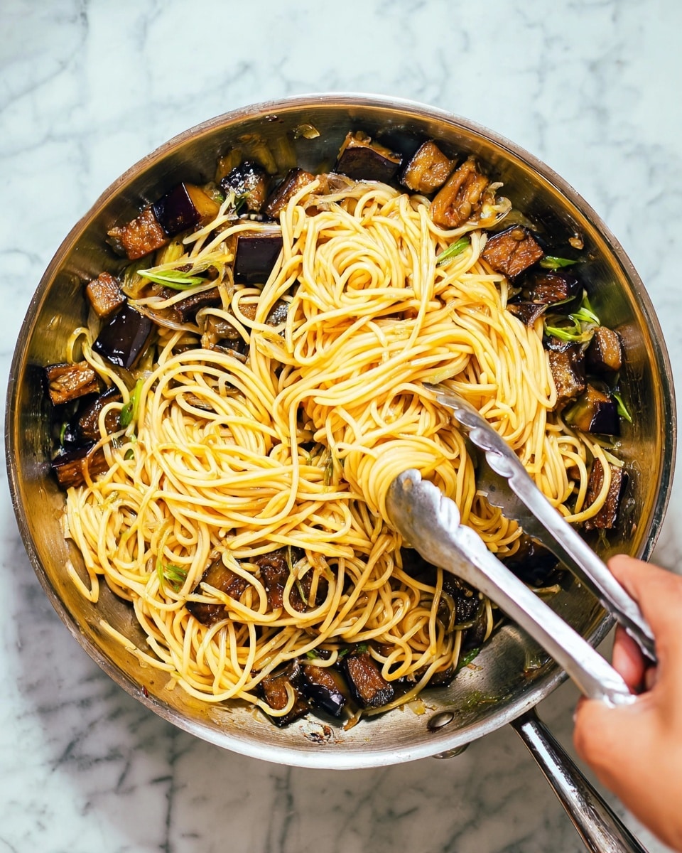 The image shows a metal frying pan filled with yellow cooked noodles mixed with chunks of dark brown cooked eggplant. The noodles are long, smooth, and arranged loosely over the eggplant pieces. Some small pieces of green vegetable, likely sliced scallions, are scattered among the noodles and eggplant. A woman's hand is holding silver tongs that grasp some noodles in the middle of the pan. The pan is placed on a white marbled surface, with natural light brightening the scene. Photo taken with an iphone --ar 4:5 --v 7