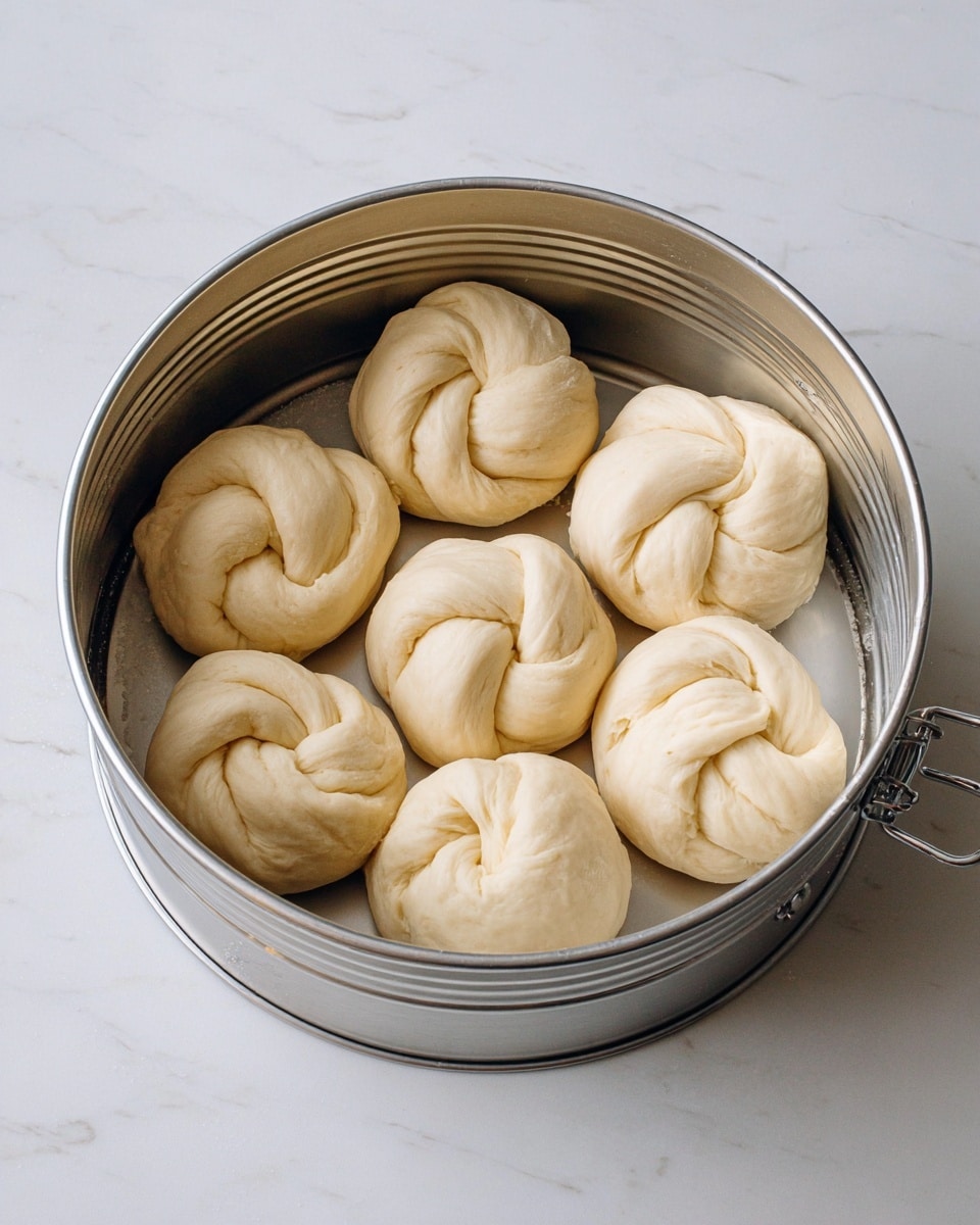 The image shows seven pieces of dough shaped into round knots placed evenly inside a silver metal baking pan. Each dough knot has smooth, light beige texture with soft creases and folds visible on the surface. The baking pan is open and shiny, with some light scratches and a latch on the side. The pan sits on a white marbled surface that is bright and clean, providing a soft background to the dough knots. photo taken with an iphone --ar 4:5 --v 7