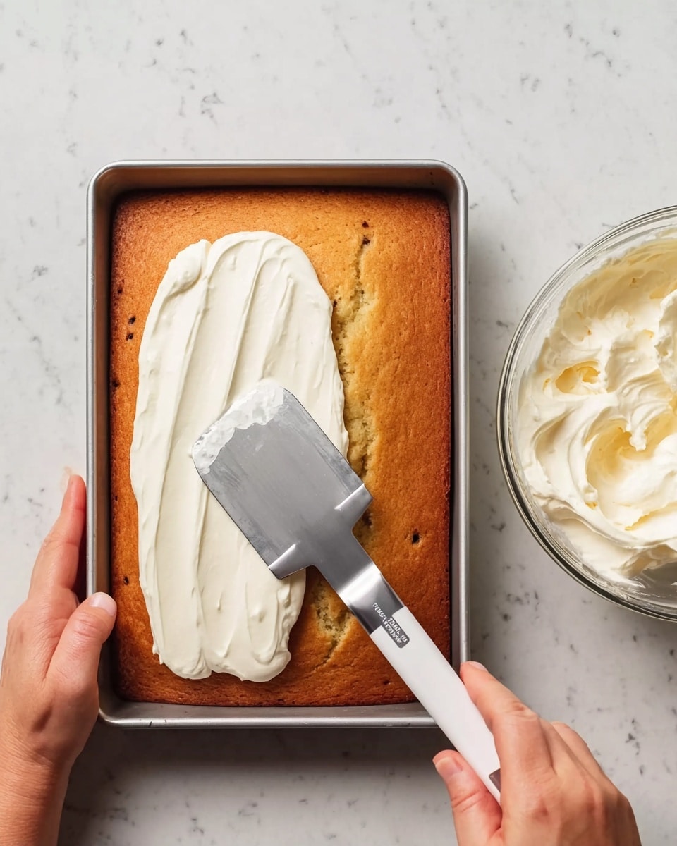 A rectangular golden brown cake with small darker spots is in a metal baking pan placed on a white marbled surface. On the left side of the cake, thick white frosting is spread in uneven layers, with a shiny metal spatula held by a woman's hand smoothing the frosting over the cake. Another woman's hand is steadying the pan from the bottom left corner. To the right of the pan, there is a clear glass bowl filled with white frosting. Photo taken with an iphone --ar 4:5 --v 7