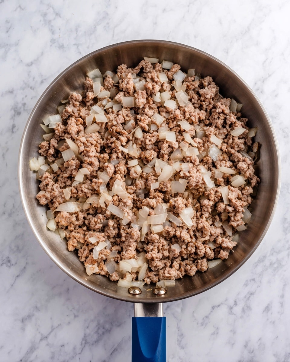 A stainless steel pan filled with cooked ground meat and diced onions evenly mixed together. The ground meat has a crumbly texture in light brown tones with some pieces slightly darker, while the diced onions are translucent white and spread throughout the pan. The pan has a blue handle and sits on a white marbled surface, showing a top-down view of the contents. Photo taken with an iphone --ar 4:5 --v 7