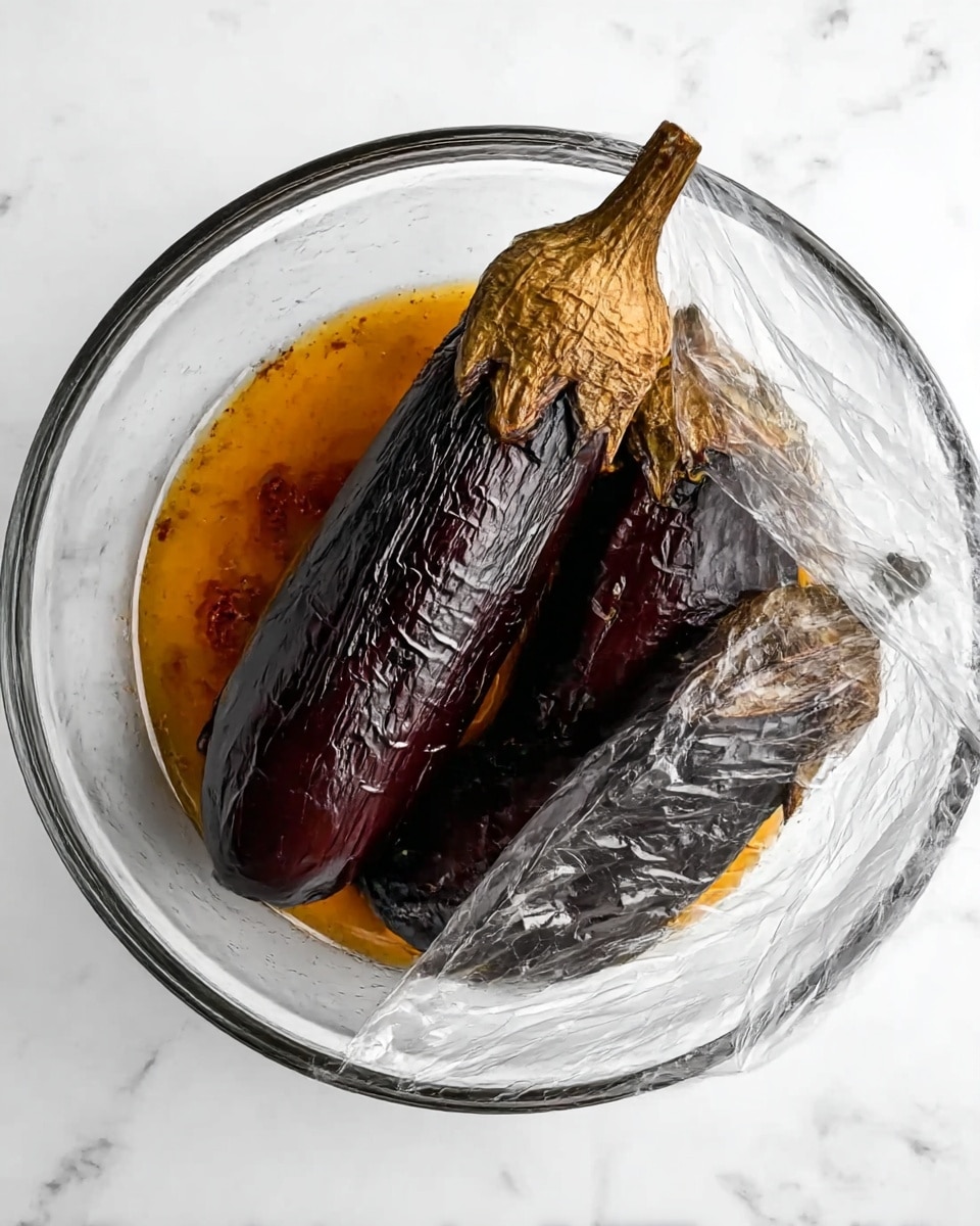 The image shows two eggplants in a glass bowl on a white marbled surface. The eggplants are dark purple with blackened, wrinkled skin, showing they have been roasted. The tops of the eggplants are light brown and dry. There is some orange-brown liquid at the bottom of the bowl. A transparent plastic wrap is partially covering the bowl, gathered to one side. Photo taken with an iphone --ar 4:5 --v 7