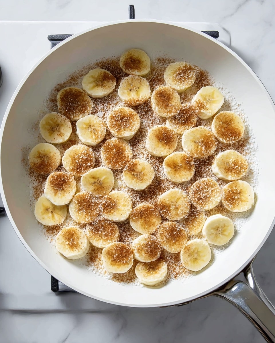 The image shows a white pan on a white marbled surface filled with about 30 round banana slices arranged in a single layer. Each banana slice is topped with a light dusting of brown cinnamon powder and white sugar crystals, creating a contrast in color and texture on the soft, yellowish banana pieces. The edges of the pan and burner handles are visible, adding a metallic shine to the setting. photo taken with an iphone --ar 4:5 --v 7