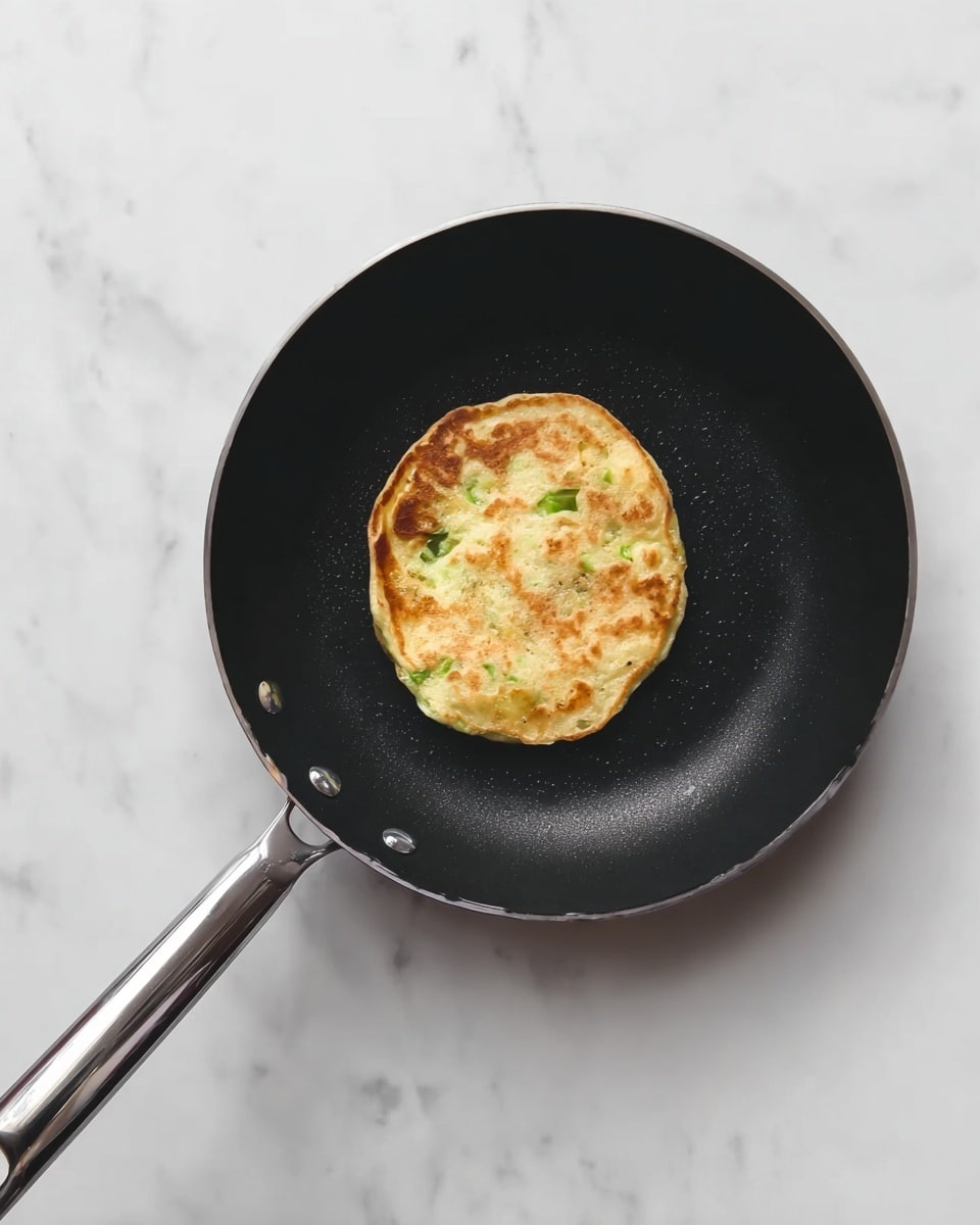 A small, round pancake with light golden brown spots cooking in the center of a black frying pan with a shiny silver handle. The pancake has a slightly uneven edge and green bits of vegetables visible inside. The frying pan is placed on a white marbled surface. photo taken with an iphone --ar 4:5 --v 7