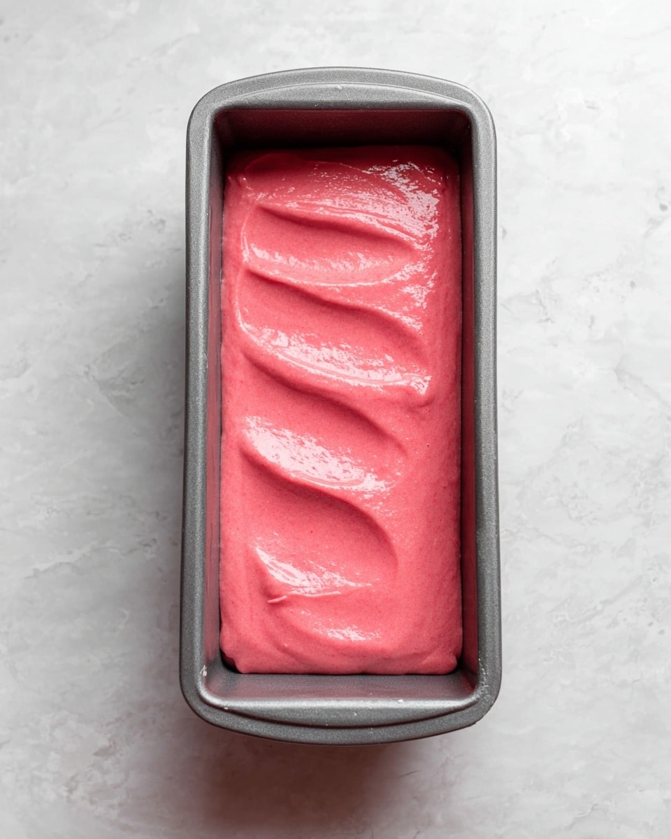 A gray metal loaf pan holds a single layer of thick, bright pink batter with a shiny, smooth surface and a few gentle waves running across it. The pan is placed on a white marbled surface, creating a clean and simple background. photo taken with an iphone --ar 4:5 --v 7