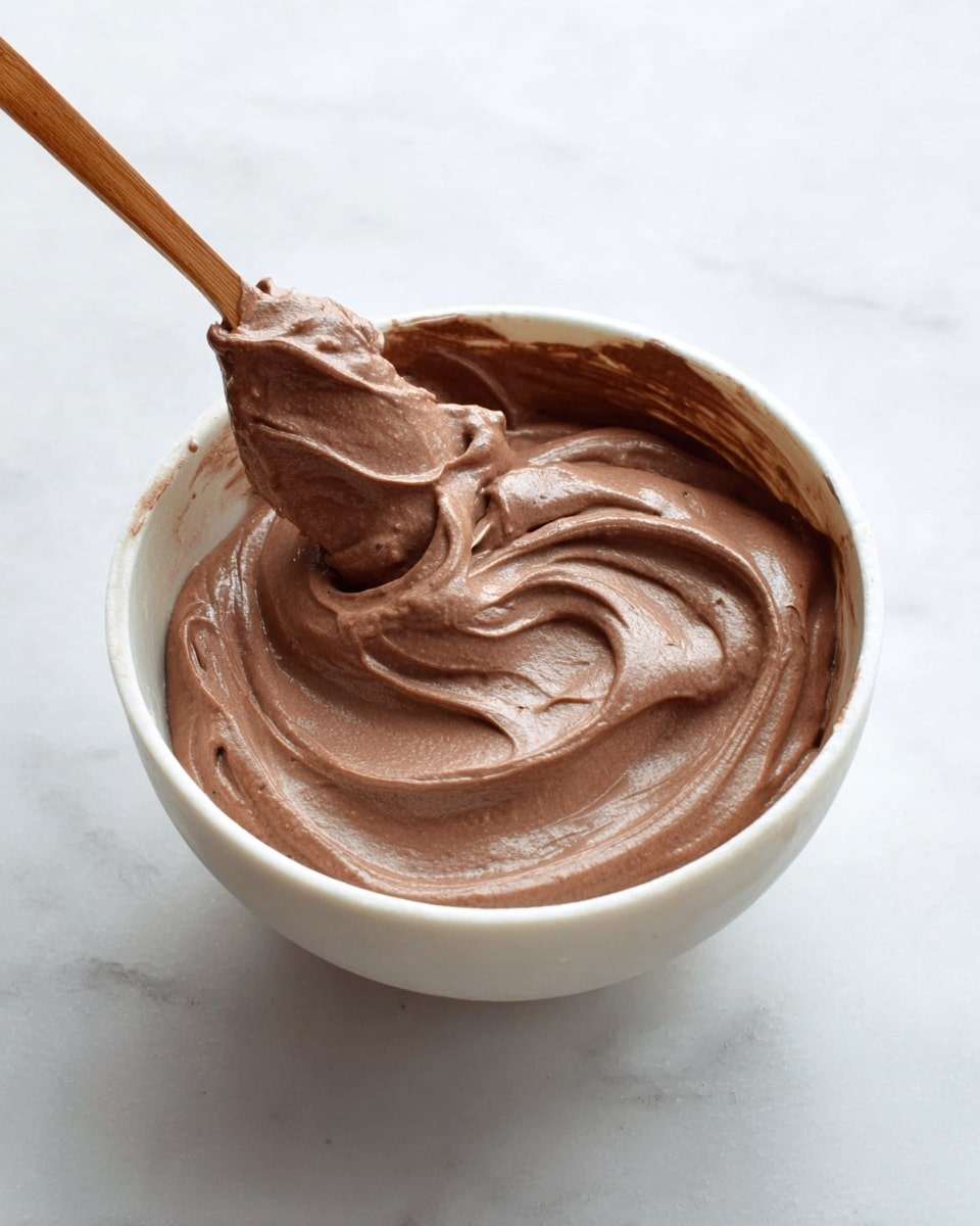 A white bowl filled with thick, creamy chocolate mousse with a smooth texture and swirled peaks, sitting on a white marbled surface. A brown spoon is dipping into the mousse, showing its rich and soft consistency. The mousse clings lightly to the sides of the bowl, creating a rich brown contrast against the white bowl. photo taken with an iphone --ar 4:5 --v 7