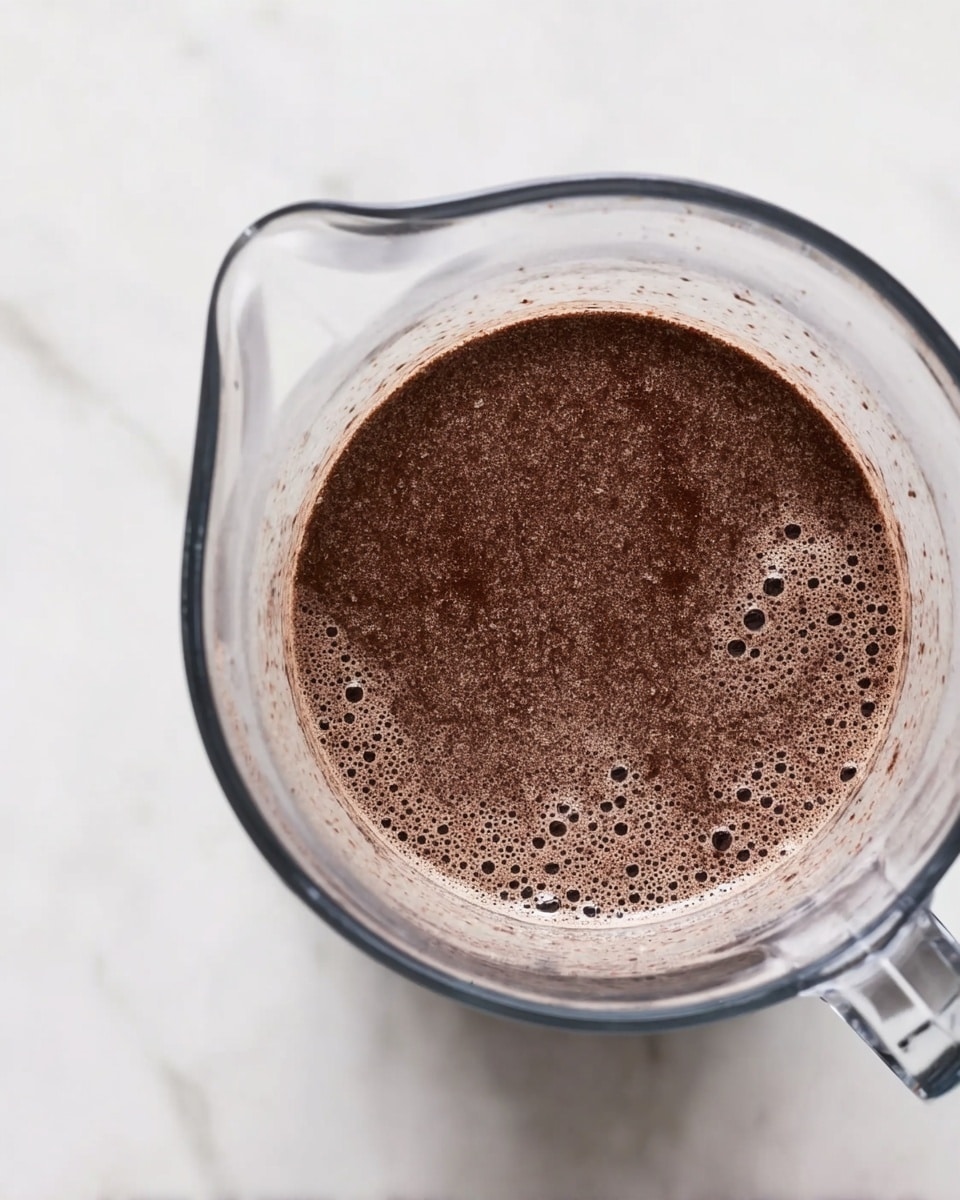 A clear glass blender jar sits on a white marbled surface, filled with a thick, dark brown mixture that has a slightly frothy texture with small bubbles on its surface. The blender jar’s handle and spout are visible on the top right and left sides, showing the smooth, transparent glass edges. The mixture fills the jar almost to the top, creating a uniform dark brown layer with tiny bits of texture throughout. photo taken with an iphone --ar 4:5 --v 7