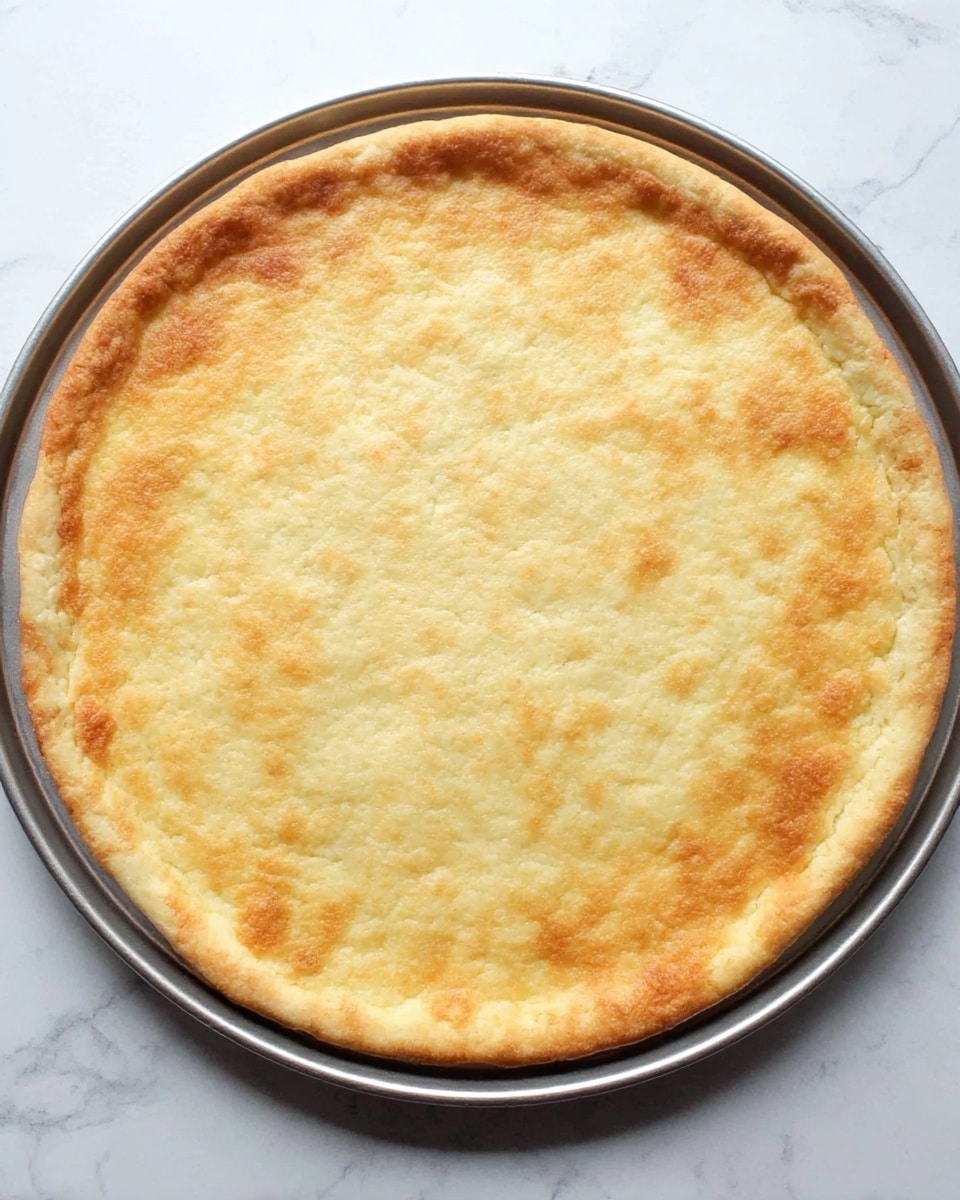 A single layer of golden-baked pizza crust with a slightly uneven round shape, showing light golden-brown edges and a soft, slightly textured surface in the center, placed on a round metal pan that rests on a white marbled background photo taken with an iphone --ar 4:5 --v 7