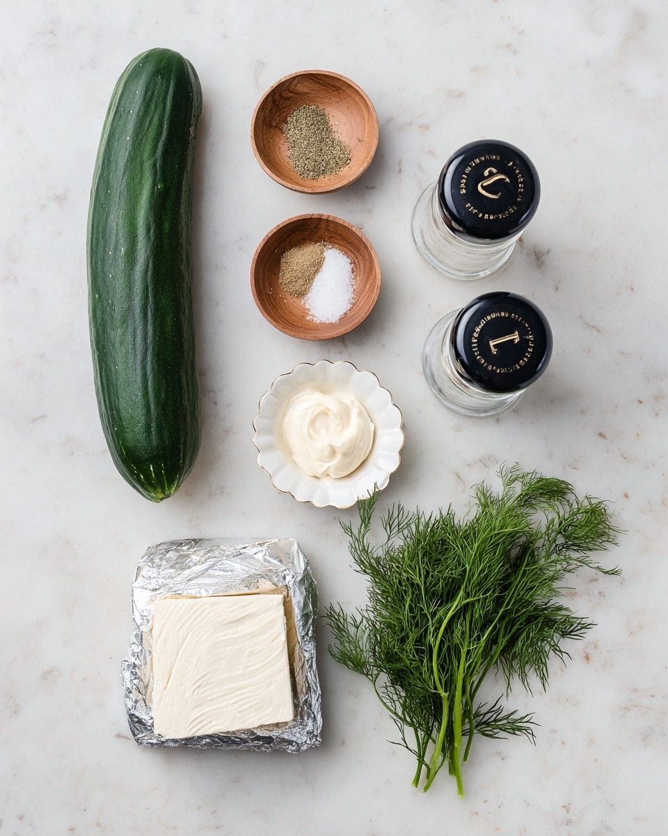 The image shows ingredients arranged neatly on a white marbled surface, including a long dark green cucumber on the left, a silver wrapped block of cream cheese below it, and a small bunch of fresh green dill to the right. Above these, there are two small wooden bowls with fine powdered spices, a small white scalloped dish with a dollop of mayonnaise, and two clear salt and pepper shakers with black tops labeled