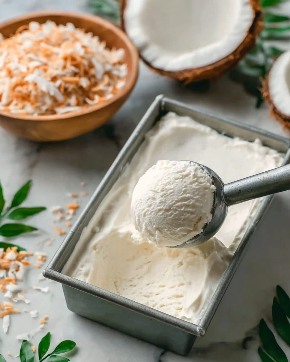 The image shows a full metal rectangular container filled with smooth white ice cream. A metal ice cream scoop is lifting a round, textured scoop of the creamy, white ice cream from the container, slightly above the surface. In the background, there is a white bowl filled with toasted shredded coconut and a halved coconut shell with white coconut flesh inside, both resting on a white marbled surface with some green leaves scattered nearby. photo taken with an iphone --ar 4:5 --v 7