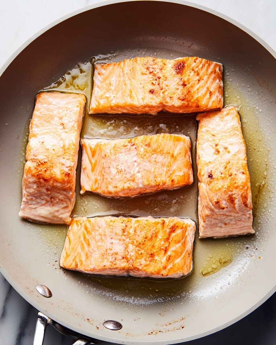 The image shows four pieces of salmon cooking in a round, light gray non-stick pan. Each salmon piece is rectangular with a golden-brown sear on the top surface, and the sides have a light pink color with some visible texture from the fish. The pan has some oil spread unevenly around the pieces, giving a shiny and slightly oily look. The pan's edges and handle are visible, and the background is a white marbled surface. photo taken with an iphone --ar 4:5 --v 7