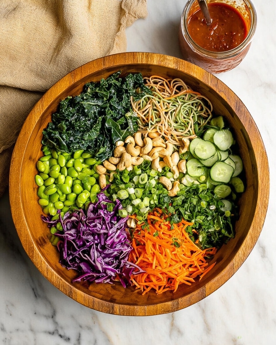 A wooden bowl filled with eight sections of fresh ingredients arranged in a circular pattern: bright green chopped kale on the top left; pale brown thin noodles mixed with dark green leafy greens below it; deep purple shredded cabbage at the bottom left; light beige cashew nuts next to the cabbage; bright green edamame beans above the cashews; bright orange thin carrot sticks on the right; fresh green sliced cucumber above the carrots; and chopped green herbs with some sliced green onions in the center. The bowl is set on a white marbled surface with a beige cloth napkin in the top left corner and a small jar of reddish-brown dressing in the top right corner. photo taken with an iphone --ar 4:5 --v 7