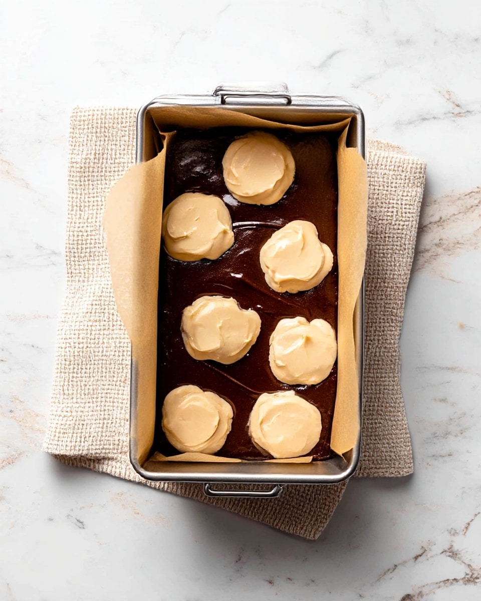 A rectangular metal baking pan lined with parchment paper holds a dark brown chocolate batter spread evenly as the bottom layer. On top, there are eight dollops of a light tan batter spaced out in two rows, their smooth and creamy texture contrasting with the darker base underneath. The pan sits on a beige textured cloth, all placed on a white marbled surface. Photo taken with an iphone --ar 4:5 --v 7