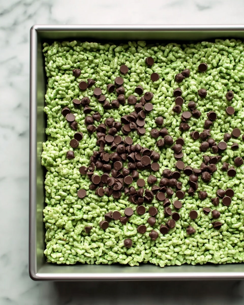 A square metal pan filled with a single thick layer of green puffed rice treats. The texture is bumpy and crispy, with many puffed rice pieces closely packed together. Scattered on top are small, round dark brown chocolate chips spread unevenly across the green surface. The pan sits on a white marbled surface. Photo taken with an iphone --ar 4:5 --v 7