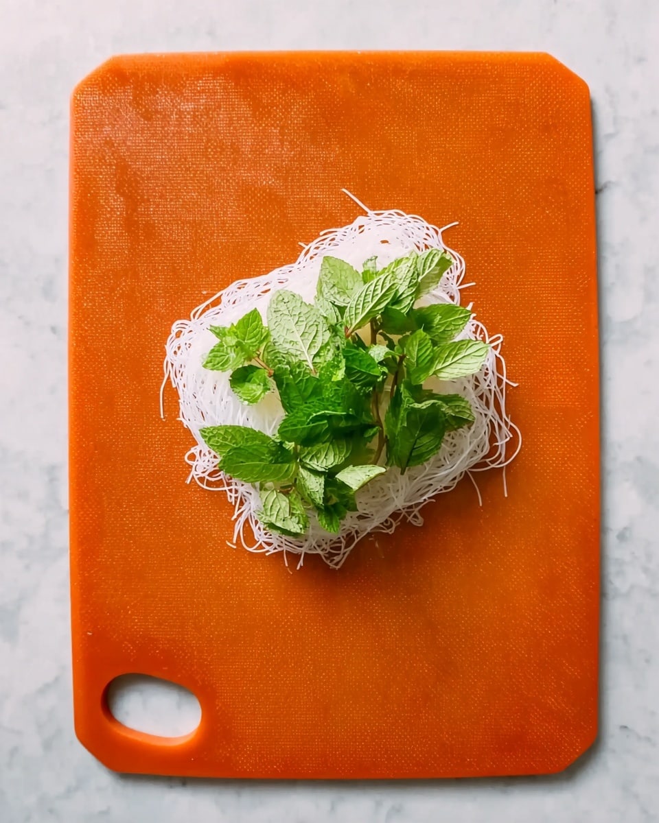 A single transparent rice paper sheet is placed on an orange cutting board with a small round hole on the bottom left. On the middle of the rice paper, there is a neat pile of thin white rice noodles forming the base layer. On top of the noodles, there are fresh green mint leaves spread evenly, adding a leafy texture and bright green color. The cutting board is on a white marbled surface. photo taken with an iphone --ar 4:5 --v 7