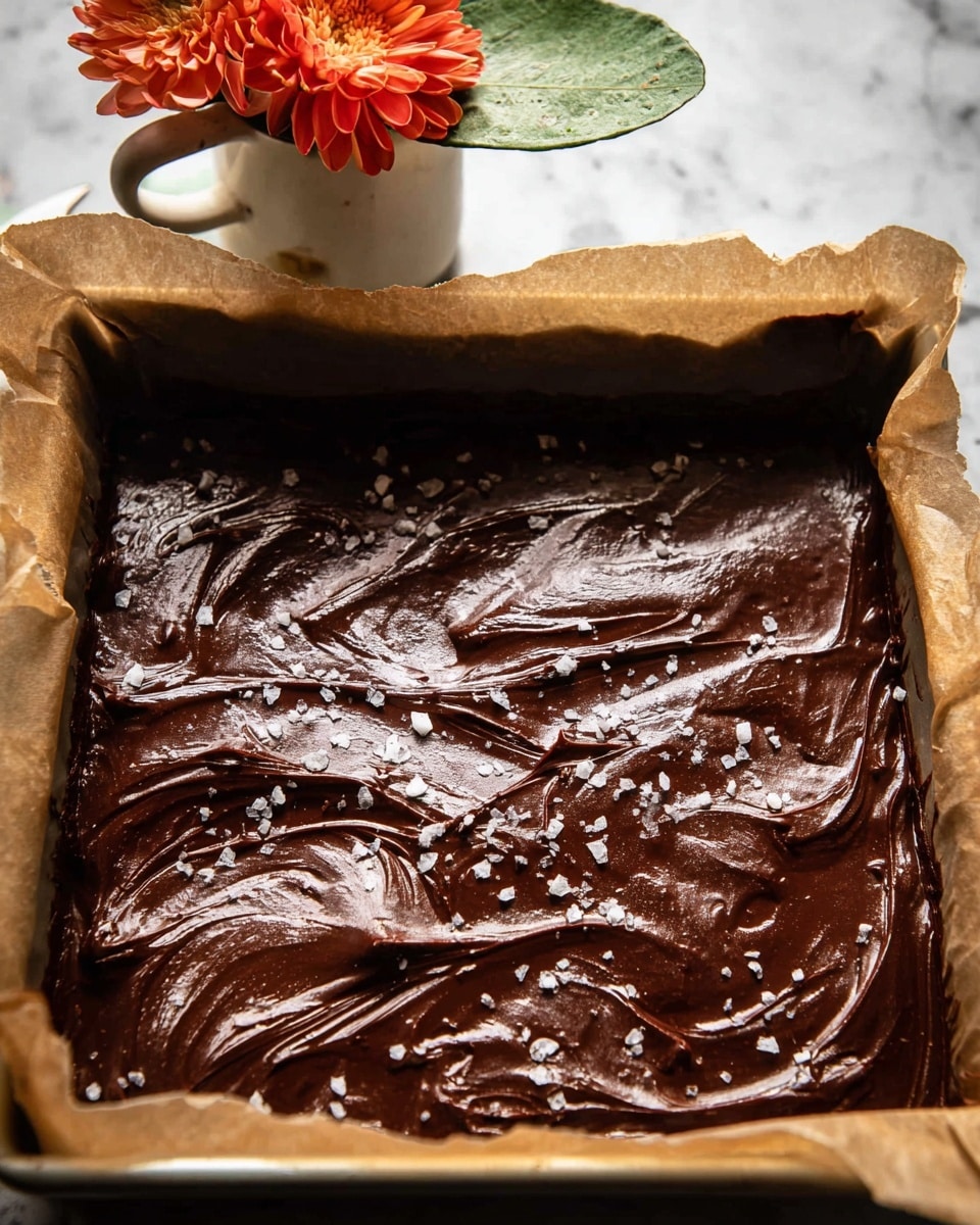 The image shows a close-up of a thick layer of dark brown chocolate spread evenly in a baking pan lined with parchment paper. The chocolate surface has visible swirls and waves with a shiny, smooth texture. On top, there are flakes of coarse salt scattered irregularly, adding a contrasting white color to the chocolate. In the background, there is a small white ceramic cup holding an orange flower and a green leaf, resting on a white marbled surface. The image is warm and detailed with a cozy atmosphere. photo taken with an iphone --ar 4:5 --v 7
