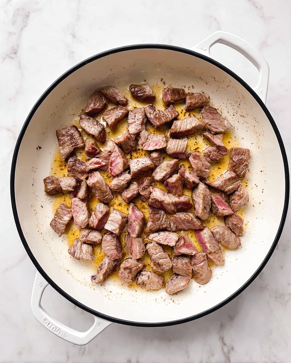 Inside a large white pan with a black rim and white handles, there are many small pieces of browned meat being cooked. The meat pieces are spread out evenly, showing a mix of brown seared sides, some pinkish edges, and a little bit of juice and oil pooling in the pan underneath them. The pan sits on a white marbled surface. photo taken with an iphone --ar 4:5 --v 7