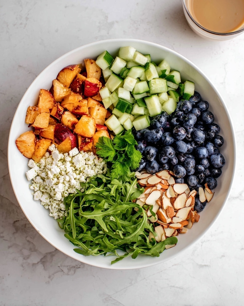 A white bowl sits on a white marbled surface, filled with five distinct sections of food arranged in a circular pattern. Starting from the top left, there are small, orange-red chunks of a fruit, followed by light green cucumber pieces with darker green skin cut into small cubes on the top right. Below the cucumber, there are many dark blue blueberries packed closely together. To the bottom right, there are light brown toasted almond slivers, next to a bright green leafy bed of arugula that fills the bottom left. On the left side, above the arugula, there is a white crumbly layer of cheese. A small sprig of fresh green herbs sits near the center where all layers meet. In the top right corner of the image, a glass cup with a light brown drink is slightly visible. photo taken with an iphone --ar 4:5 --v 7