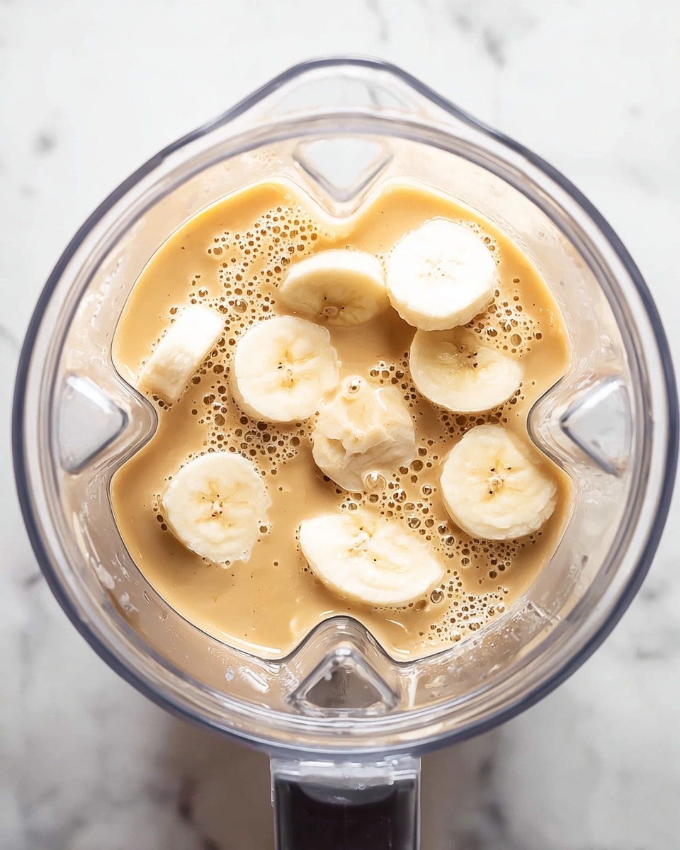 A clear blender container is viewed from above, showing a mix of light brown creamy liquid and round slices of pale yellow bananas floating inside. The bananas vary in thickness, some slices are whole while others are partial, revealing their soft texture. Tiny bubbles dot the surface of the liquid, giving it a fresh and smooth look. The blender container sits on a white marbled surface, adding a clean background to the image. Photo taken with an iphone --ar 4:5 --v 7