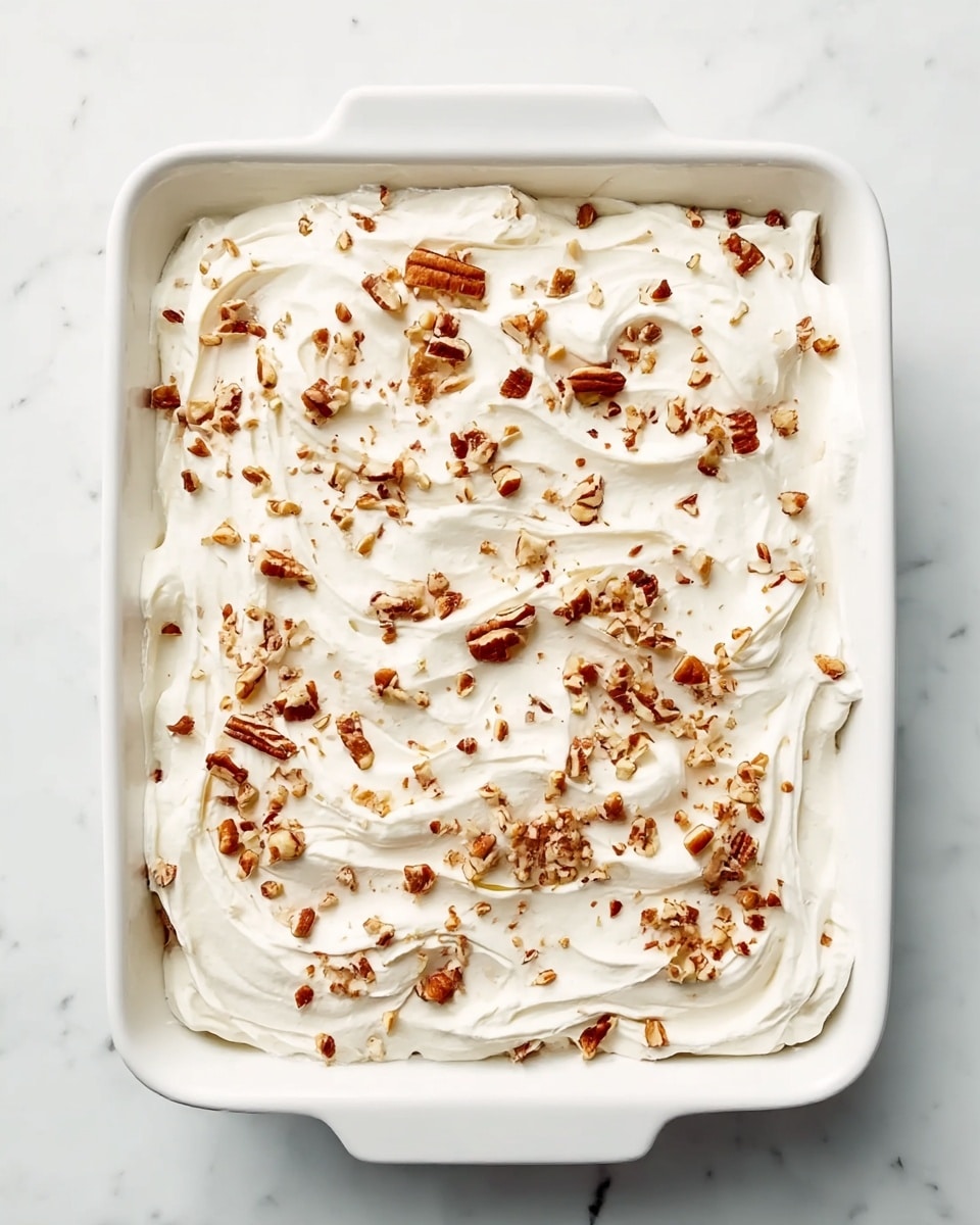 A white rectangular dish filled with a thick layer of creamy white frosting, spread smoothly with visible swirls and soft peaks across the whole surface. On top, there is a generous sprinkle of chopped brown nuts scattered evenly, adding texture and color contrast against the white frosting. The dish sits on a white marbled surface, highlighting the freshness and simplicity of the dessert. Photo taken with an iphone --ar 4:5 --v 7