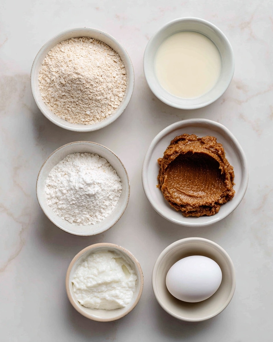The image shows six small white bowls and one white egg placed on a white marbled surface. The top left bowl contains fine, light beige oat flour. To its right is a bowl filled with light cream-colored milk. Below the oat flour is a white bowl holding thick, brown date paste with a smooth texture and some brown smudges on the edges. Next to the date paste is a single white egg. Below the egg is another white bowl filled with solid white coconut oil, which has a soft, creamy texture. At the bottom left is a white bowl containing white baking powder. Each ingredient is neatly placed with labels above them. Photo taken with an iphone --ar 4:5 --v 7