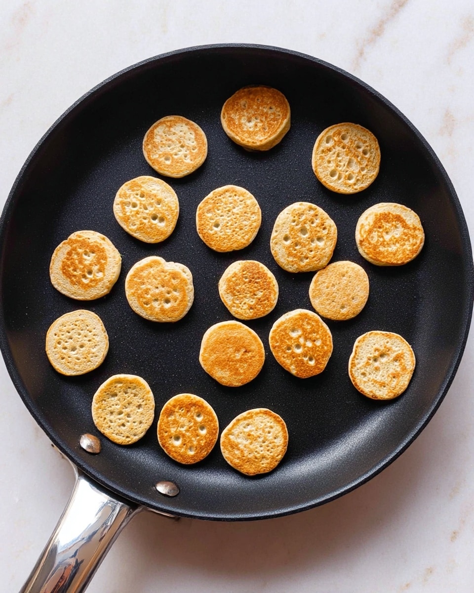 A black frying pan contains 18 small, round mini pancakes in different golden brown shades; some are light tan while others have a deeper toasted color. The pancakes vary slightly in size and have small bubbles and porous textures on top. They are spread out in an irregular pattern across the pan, with a few showing smooth edges and others having rough, lumpy sides. The pan handle is silver and shiny, and the pan sits on a white marbled surface. Photo taken with an iphone --ar 4:5 --v 7