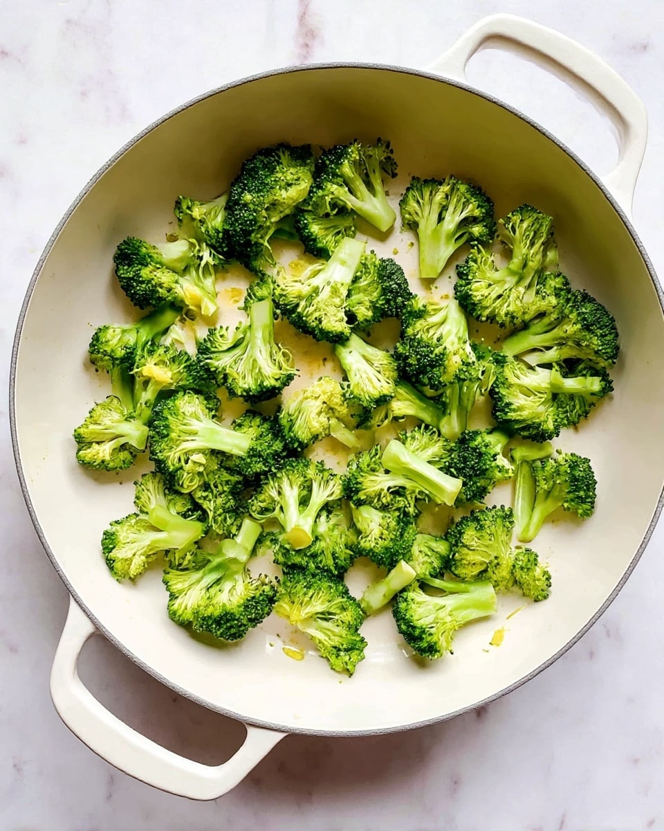 Inside a white pan with two handles, there are many small bright green broccoli florets spread evenly. The broccoli shows detail in the texture, with tiny buds and stalks visible, and some light yellow hints where they have been cooked a little. The pan sits on a white marbled surface. photo taken with an iphone --ar 4:5 --v 7