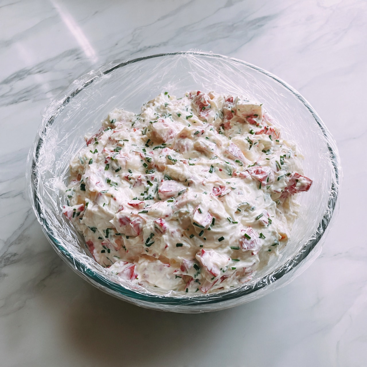 A clear glass bowl sits on a white marbled surface, filled with a mixture of chopped white and pink pieces, coated evenly in a thick, creamy white sauce with small green herb bits spread throughout. The bowl is covered loosely with a clear plastic wrap that slightly crinkles around the edges. The overall look is fresh and creamy with a soft, smooth texture. photo taken with an iphone --ar 4:5 --v 7