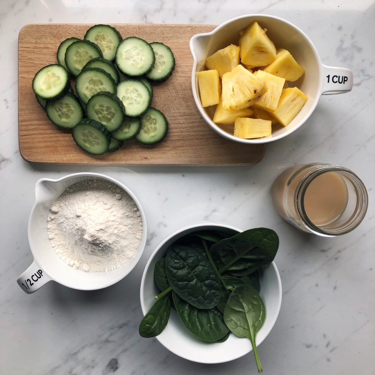 The image shows a top view of ingredients arranged on a white marbled surface. At the top left, several green cucumber slices with dark edges lie in a small pile on a wooden cutting board. To the right of the board, a white measuring cup labeled