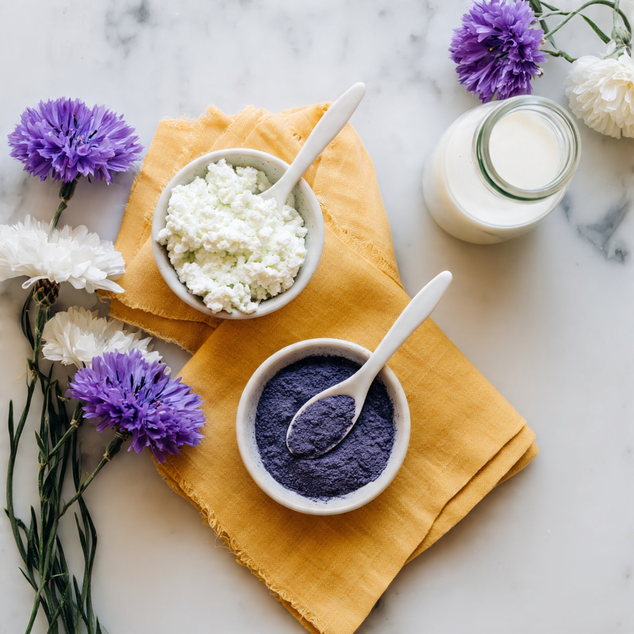 The image shows two small white dishes placed on a folded mustard yellow napkin over a white marbled surface. The top dish is filled with a fluffy white substance with a dark blue spoon partially inside it. The bottom dish contains a fine purple powder with a white spoon resting on top. To the top right is a clear bottle filled with white liquid, and to the top left are fresh purple and white flowers with green stems resting on the surface. Photo taken with an iphone --ar 4:5 --v 7