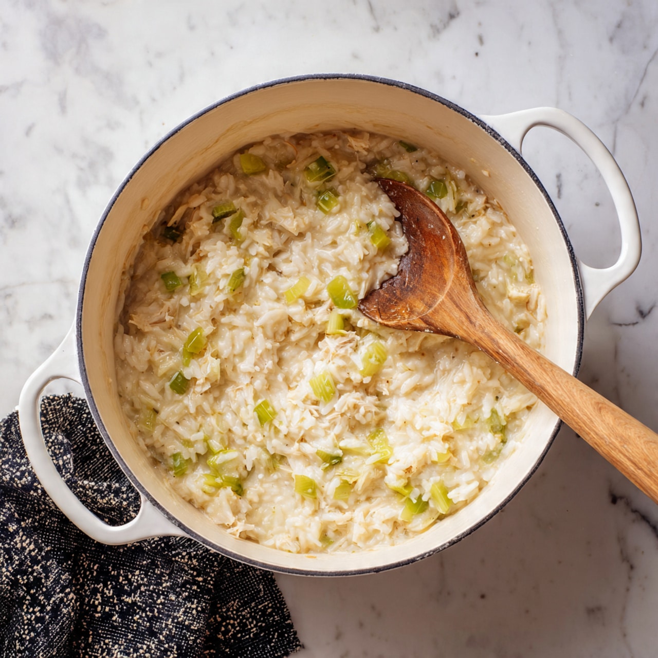 A white pot with two handles holds a creamy mixture of cooked rice and chopped green vegetables, likely celery, with a wooden spoon resting inside. The rice looks soft and thick, with a pale yellow color mixed with light green bits throughout. The pot sits on a white marbled surface with a black and white patterned cloth partially visible under the handle on the left side. The overall look is warm and homey, showing a simple, hearty food scene photo taken with an iphone --ar 4:5 --v 7