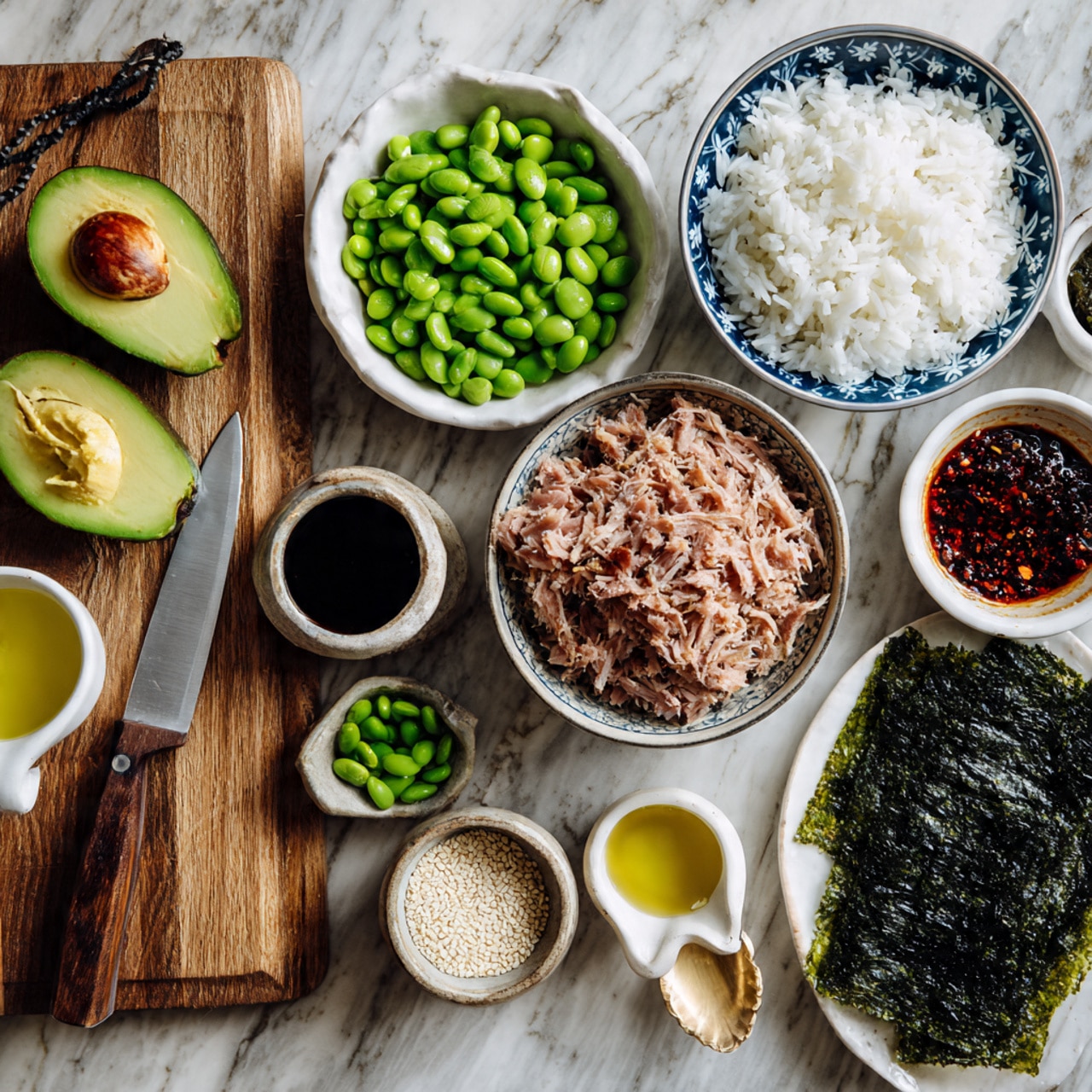 The image shows a wooden board with an avocado cut in half, one half whole and the other half diced into small green cubes, next to a knife with a silver blade and a wooden handle resting on the board. Surrounding the board are several bowls on a white marbled surface: one white bowl with green edamame, a blue patterned bowl filled with cooked white rice showing individual grains, another blue patterned bowl with shredded light pink tuna, a small beige bowl with white sesame seeds and a gold spoon inside, a small white bowl with a dark red chili oil sauce, a white cup with yellow oil, a white bowl with dark soy sauce, and a white plate holding sheets of dark green seaweed. Photo taken with an iphone --ar 4:5 --v 7