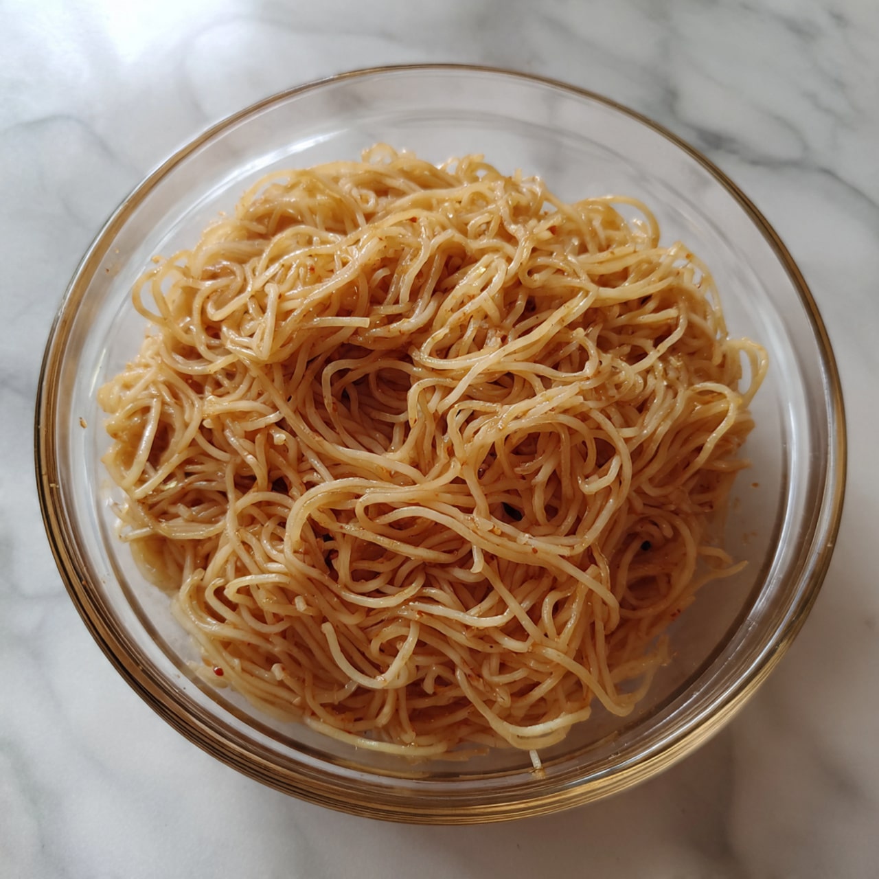 A clear glass bowl filled with thin noodles that are light brown in color, mixed with a light sauce giving some parts a slightly darker brown tint. The noodles look soft and slightly shiny, loosely piled in the bowl with some strands curling and overlapping each other. The bowl is placed on a white marbled surface, showing subtle veins and texture beneath it. The photo taken with an iphone --ar 4:5 --v 7