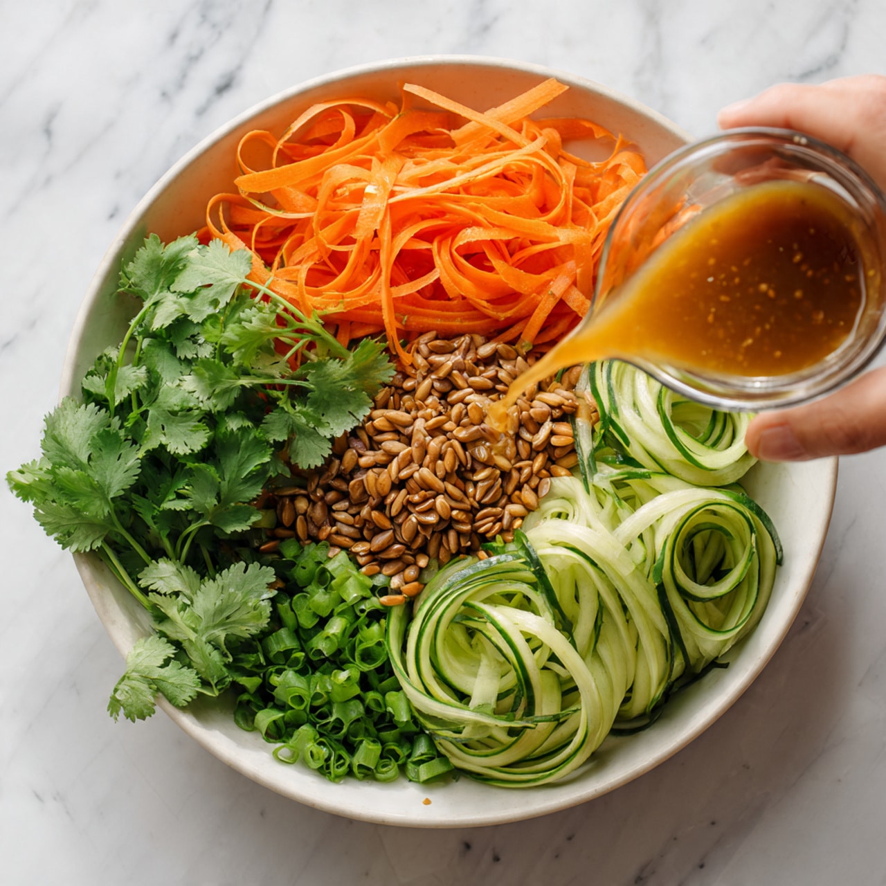 A clear round bowl sits on a white marbled surface, filled with layers of fresh ingredients. On the left side, wide orange carrot ribbons are arranged, next to a pile of light brown toasted seeds in the center. Below the seeds, a bunch of bright green cilantro leaves adds freshness. To the right, pale green cucumber ribbons curl softly. A woman's hand is holding a small clear glass cup with light brown dressing, pouring it over the salad. The scene is vibrant with fresh colors and textures, showing a healthy, crisp salad. photo taken with an iphone --ar 4:5 --v 7