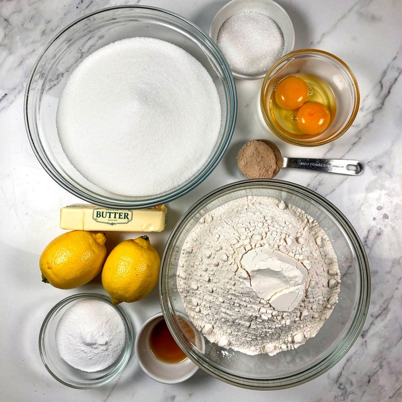 The image shows a top view of baking ingredients arranged on a white marbled surface. There are eight groups: in the top left, a large clear bowl filled with white sugar; next to it on the right, a smaller clear bowl with two bright yellow egg yolks; below this, a metal measuring spoon containing light brown powder; beneath that is a small white bowl holding a small amount of amber-colored liquid; to the left of this, a large clear bowl full of white flour; above the flour, a medium size clear bowl with white granulated sugar; to the left of the sugar, two yellow lemons lying side by side with a stick of butter wrapped in paper marked
