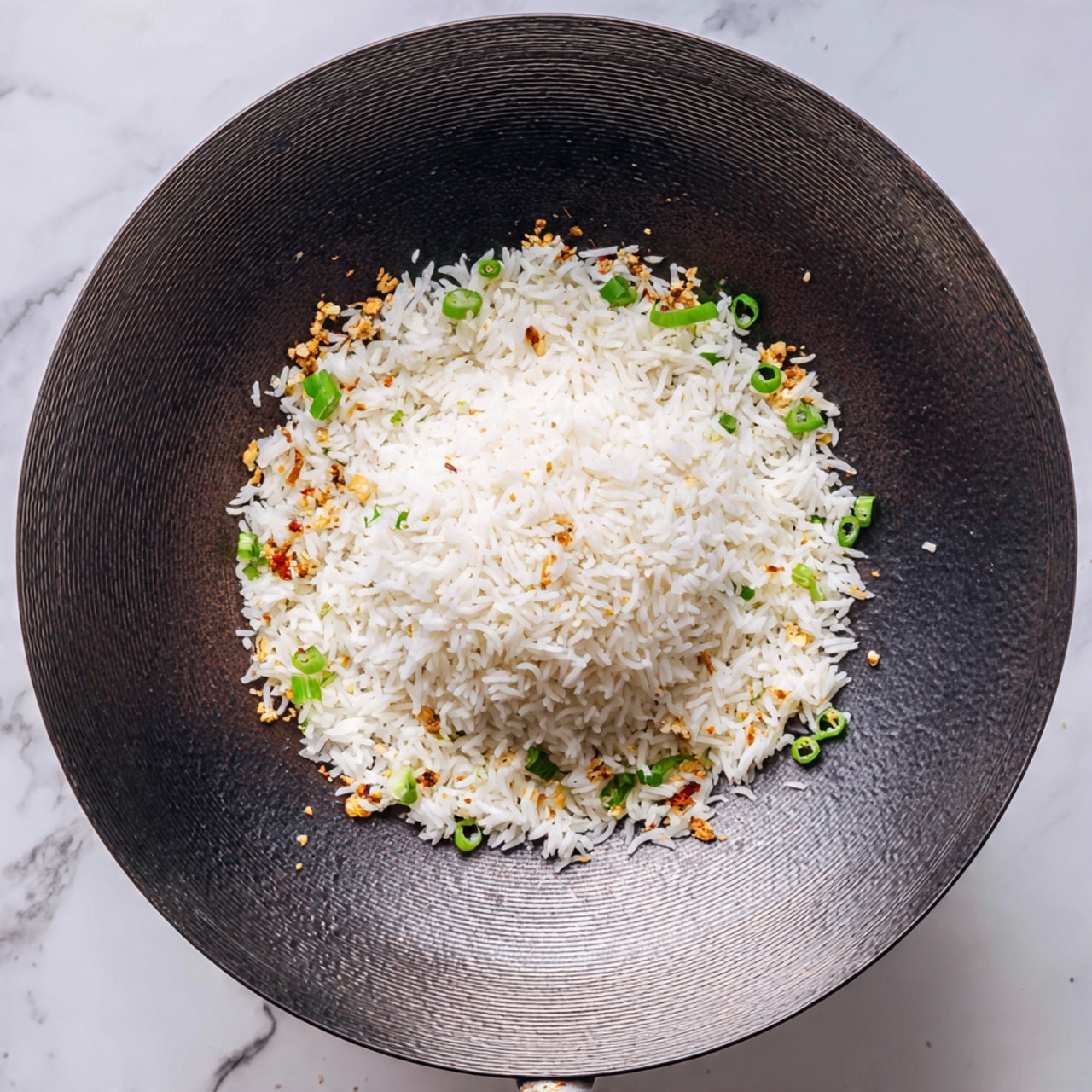 Inside a large dark wok, there is one main layer of white cooked rice centered in a mound. Surrounding the base of the rice are scattered small pieces of green onion, along with tiny bits of brown garlic or chili, adding spots of green and brown color around the white rice. The wok has a textured, shiny metal surface and is set against a white marbled background. photo taken with an iphone --ar 4:5 --v 7