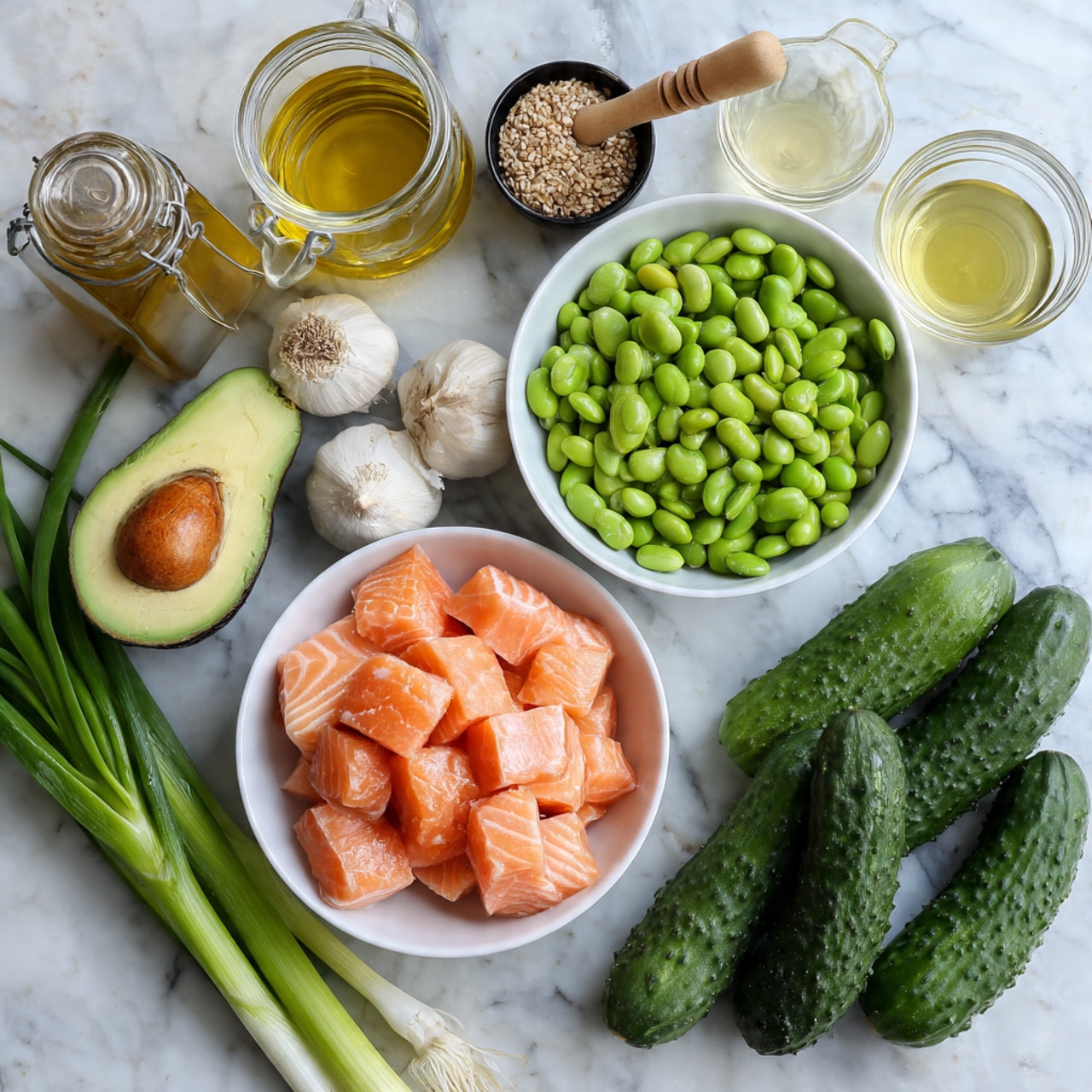 The image shows a top-down view of various fresh ingredients arranged neatly on a white marbled surface. At the center bottom, there is a white bowl filled with bright orange cubes of salmon, each piece smooth and moist in texture. To the top right of the salmon bowl, a white bowl full of bright green edamame beans, plump and shiny, sits next to a cluster of fresh green onions with long, slender green stalks and white bulbs extending toward the bottom right. To the right side, three medium-sized cucumbers with dark green bumpy skin lay side by side. Above the cucumbers, a halved avocado with a rough green outer peel and a smooth light green inside, with a large brown seed in the middle, is placed. On the left side, a small cluster of garlic cloves with pale beige papery skin and a small head rest near a set of glass containers filled with various liquids: a tall bottle of light yellow olive oil, a jar of golden honey with a honey dipper resting inside, a glass bowl with a pale clear liquid, and a small black bowl with reddish-orange sauce. Also nearby are a small white bowl with creamy white mayonnaise and several other small containers holding dark soy sauce, red crushed chili flakes, and a tiny black bowl with white salt. The overall scene is bright, colorful, and neatly organized, displaying textures from smooth fish to fresh vegetables and various sauces photo taken with an iphone --ar 4:5 --v 7