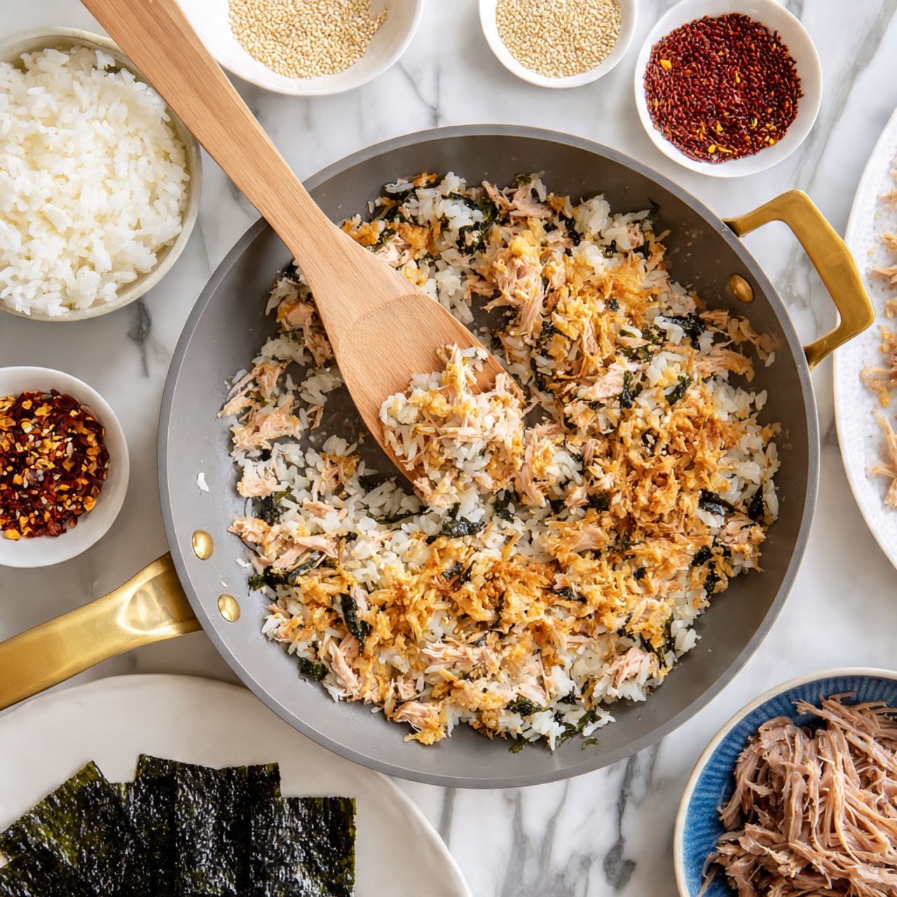 A close-up of a gray pan with gold handles filled with white and golden-brown fried rice that has small crispy bits on top and mixed throughout. A wooden spatula in the center scoops up some of the rice. Around the pan, there are small white dishes holding red chili flakes, white sesame seeds, a white bowl with white rice, a white plate with sheets of dark green seaweed, and a blue bowl with shredded brown meat. All are placed on a white marbled surface. Photo taken with an iphone --ar 4:5 --v 7