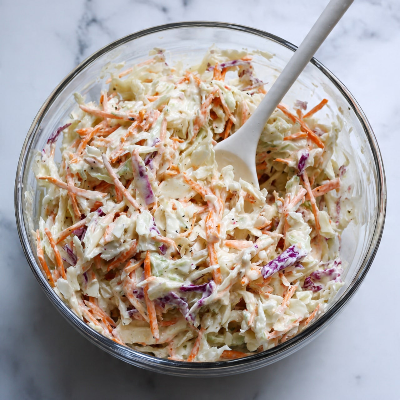 A clear glass bowl filled with shredded white cabbage, thin orange carrot strips, and some shredded purple cabbage mixed together in a creamy, light-colored dressing. The coleslaw mixture has a soft, moist texture and is being stirred with a white spoon inside the bowl. The bowl is placed on a white marbled surface. photo taken with an iphone --ar 4:5 --v 7