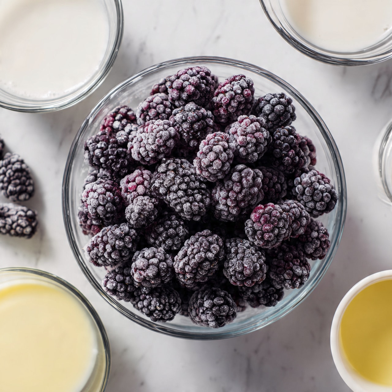 The image shows a clear glass bowl filled with dark purple frozen blackberries, with a slight icy texture covering each berry. Surrounding the bowl are clear glass measuring cups filled with white liquid cream and sugar, all placed on a white marbled surface. There is also a small white cup with a light yellow liquid, positioned near the bottom right of the frame. The overall scene is bright and clean, focusing on the ingredients ready for making a dessert or smoothie. Photo taken with an iphone --ar 4:5 --v 7